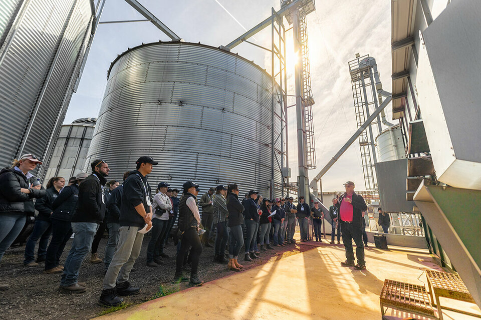 A large group of people are taken on a tour of an Alvan Blanch dryer. There are large fee silos all around